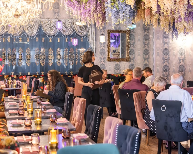 A restaurant dining room with patrons seated at tables, a server carrying menus, and floral decor.